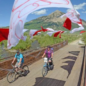 two people riding bicycles over a wooden bridge with flags flying overhead and Mt. Crested Butte in background.